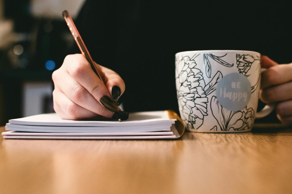 Home Person writing in a notebook with a floral ceramic mug on a wooden desk.